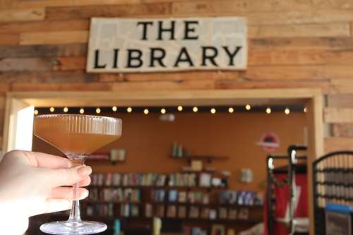 The Library Sign with books in the background and a woman's hand holding a non-alchoholic cocktail.